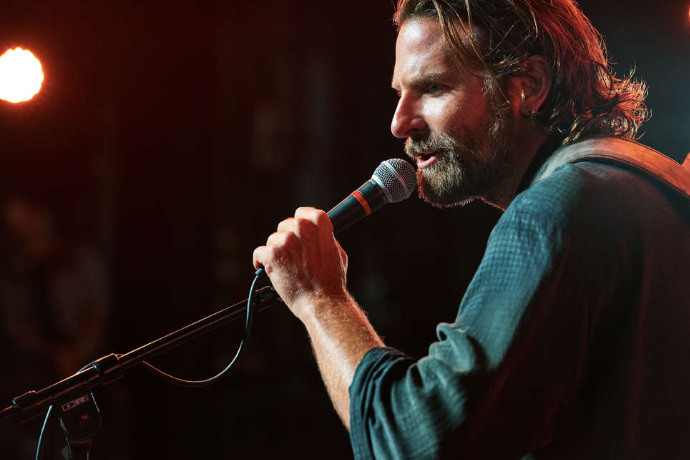 Bradley Cooper, in a blue shirt, beard, and long hair, sings into a microphone.